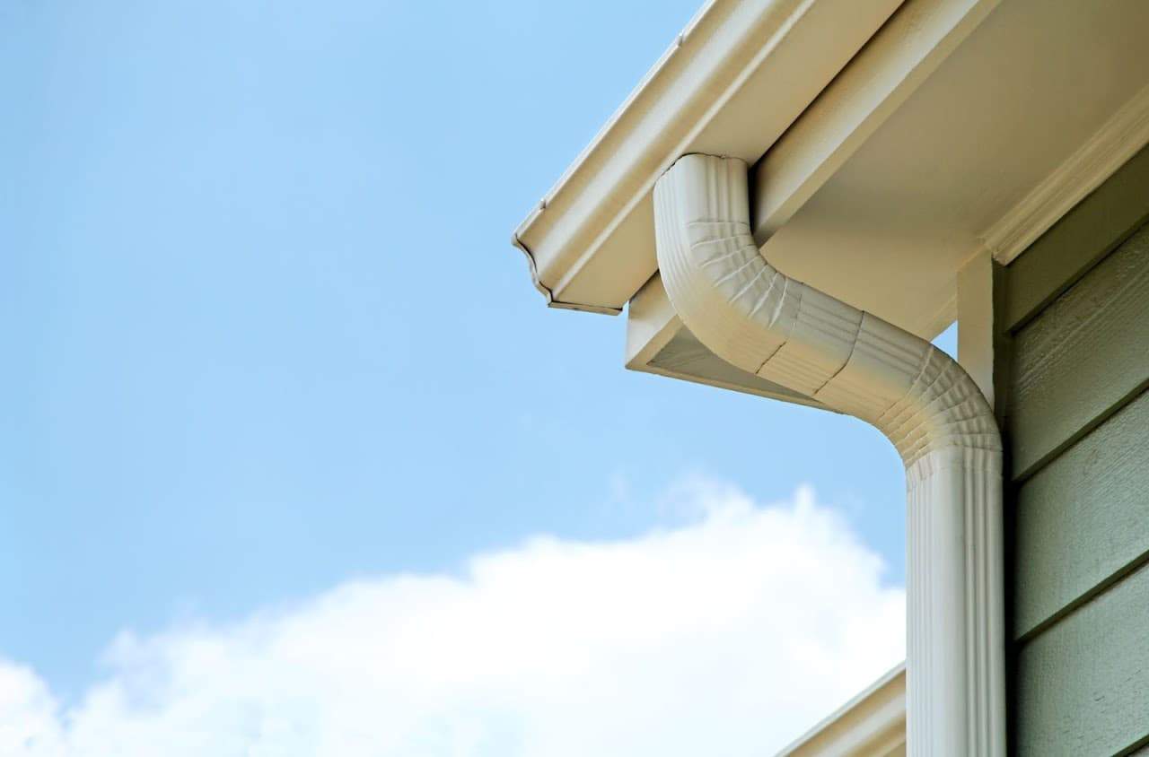 Close-up of a white rain gutter and downspout attached to the corner of a house with light green siding, set against a blue sky with some white clouds.