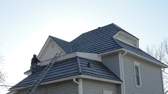 A person on a ladder is working near the roof of a light grey two-story house with blue shingles, under a clear sky.