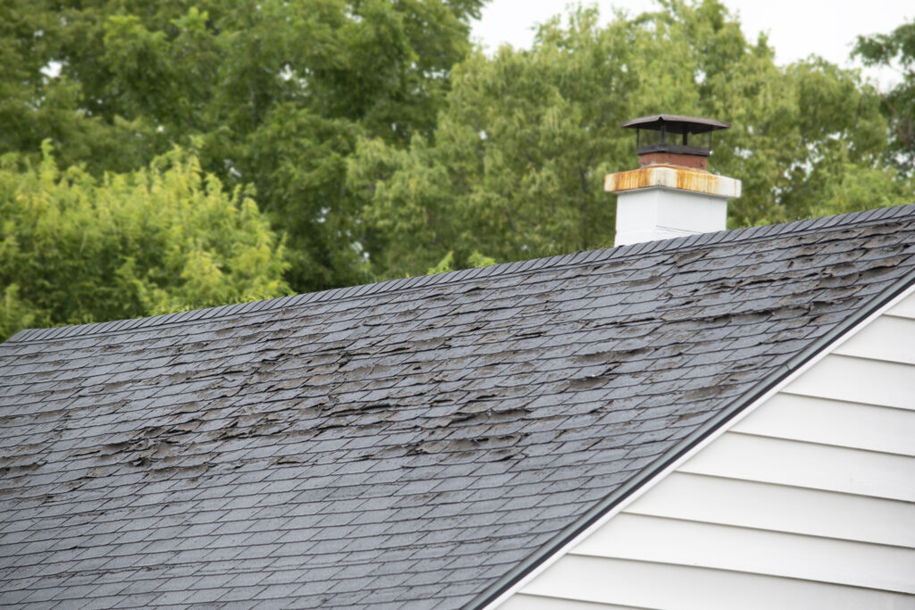 A house roof with damaged, curling asphalt shingles near a white chimney, surrounded by green trees in the background.