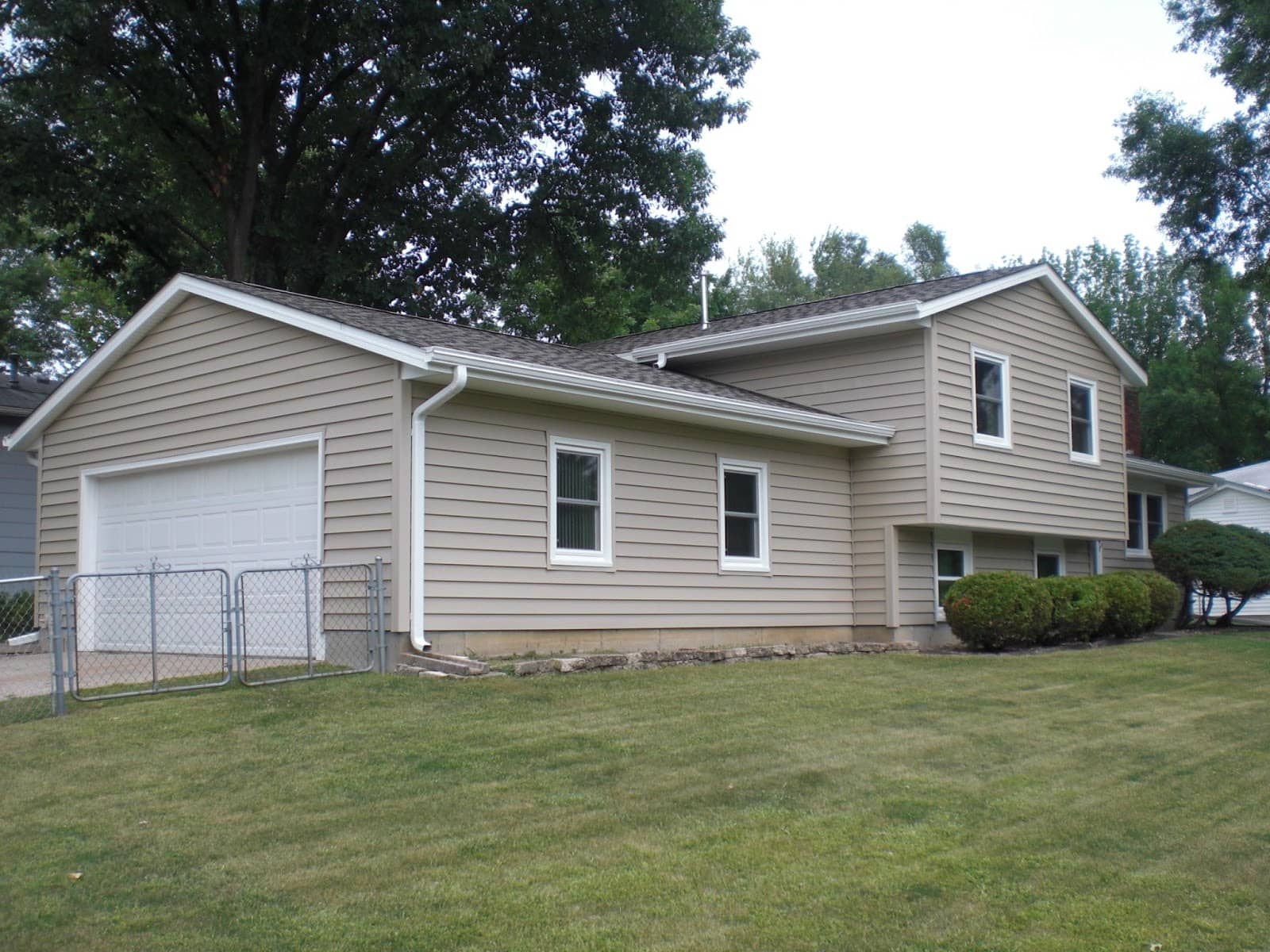 A beige split-level house with white trim, multiple windows, an attached garage, and a neatly mowed lawn, surrounded by trees and bushes.