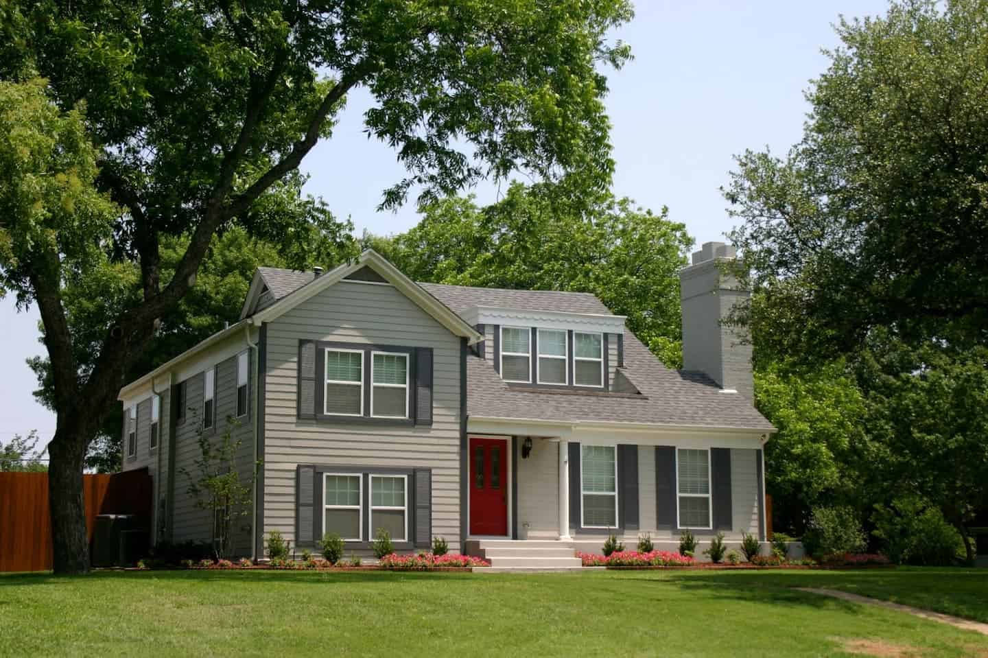 A two-story gray house with white trim and a red front door, surrounded by green grass and large trees on a sunny day. Shrubs and flowers line the front of the house.