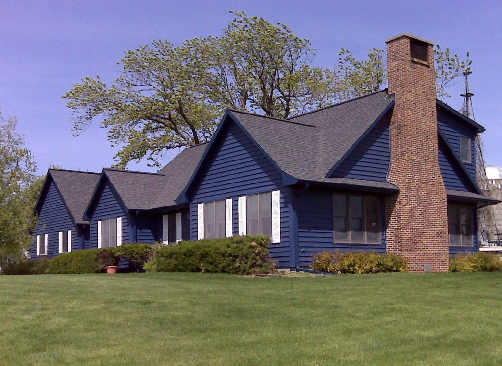 A single-story dark blue house with white-trimmed windows, a brick chimney, and a sloped roof, surrounded by green grass, bushes, and trees under a clear blue sky.