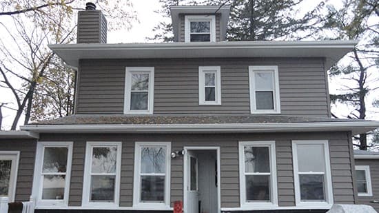 A two-story house with gray siding, white-trimmed windows, a central front door, a dormer on the roof, and trees in the background. The house has a simple rectangular shape and a small chimney.