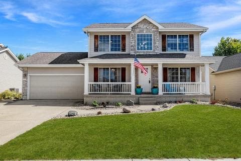 Two-story suburban house with a porch, stone and siding exterior, attached two-car garage, American flag on the porch, and a well-maintained green lawn under a blue sky.