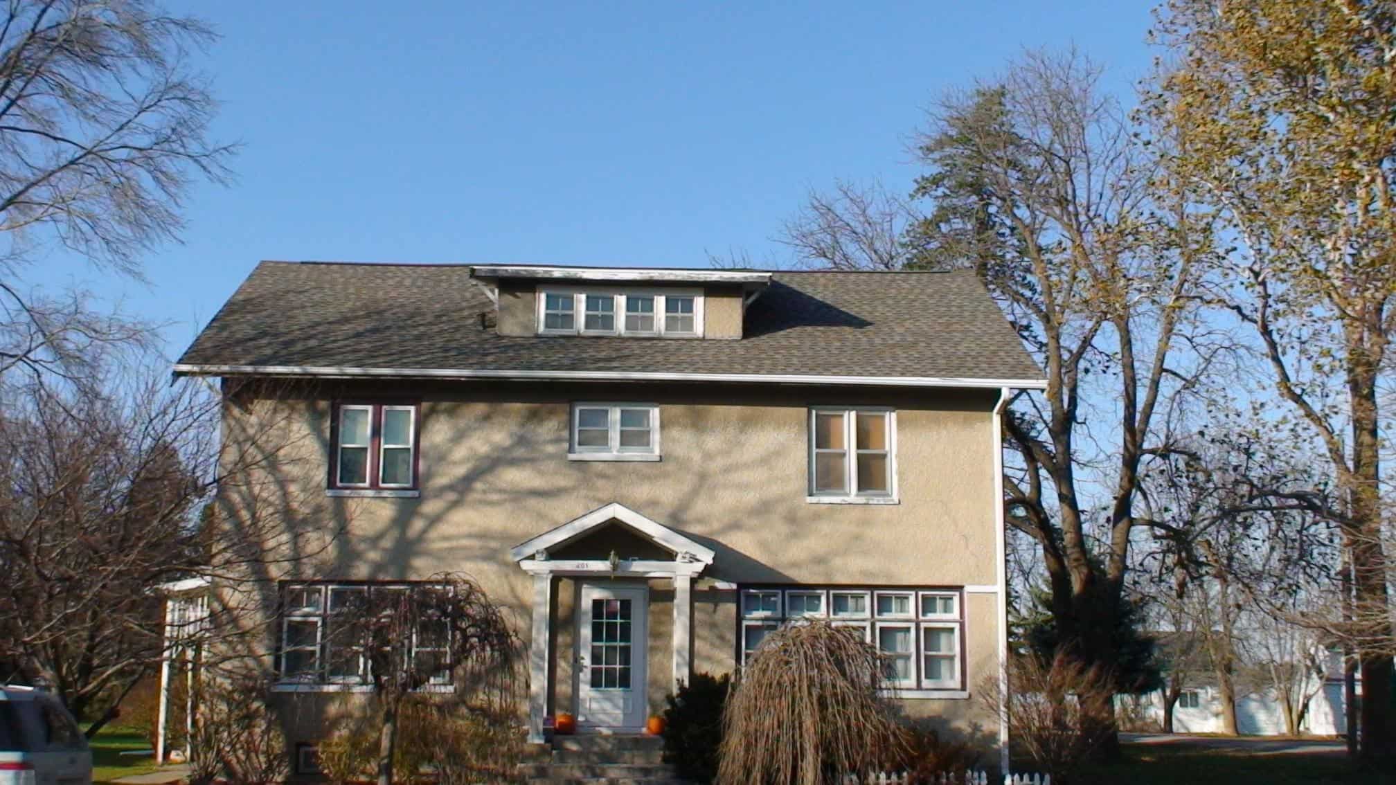 Two-story tan house with a small covered front porch, multiple rectangular windows, and a dormer on the roof, surrounded by leafless trees on a clear, sunny day.