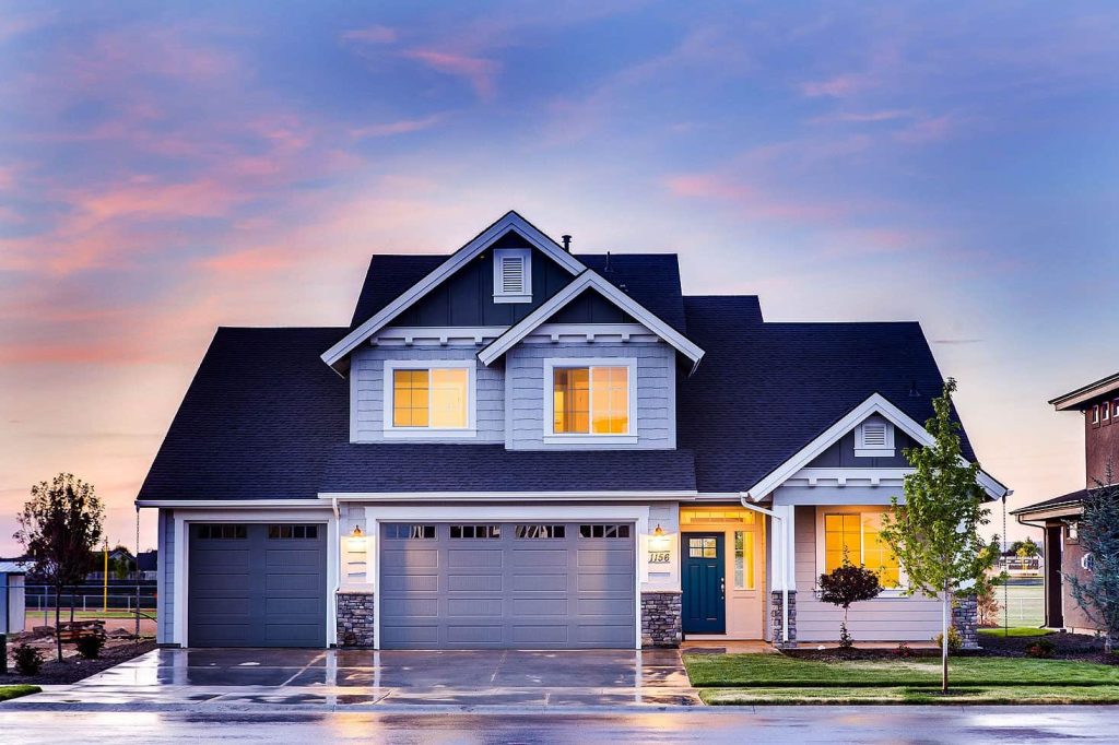 A modern two-story house with gray siding, a blue front door, three-car garage, and illuminated windows. The sky is colorful at sunset, and the driveway is wet, reflecting the house and trees.