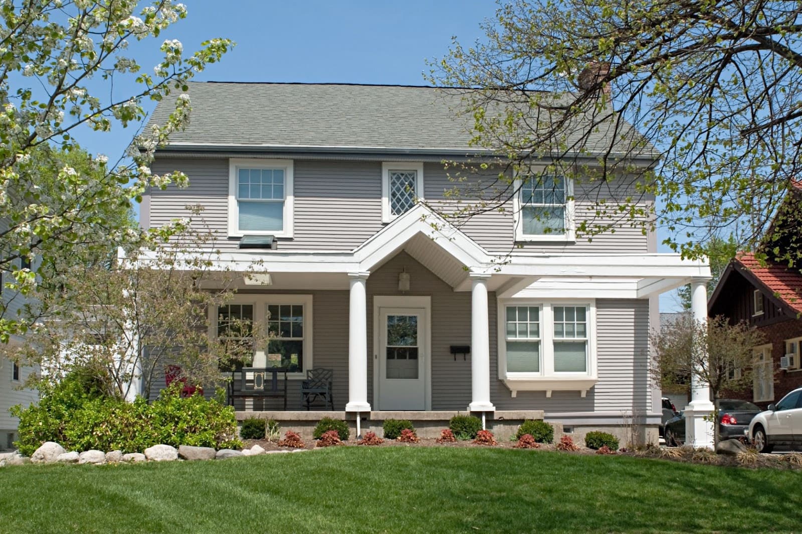 A two-story gray house with white trim, a covered front porch, and neatly landscaped yard with green grass and small bushes, under a blue sky with trees partially in view.