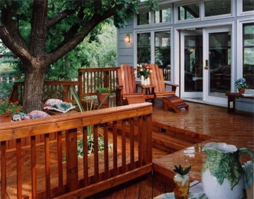 A cozy wooden deck with chairs, surrounded by a wooden railing and shaded by a large tree, leads to glass doors of a house. Potted plants and outdoor decorations are arranged around the deck.