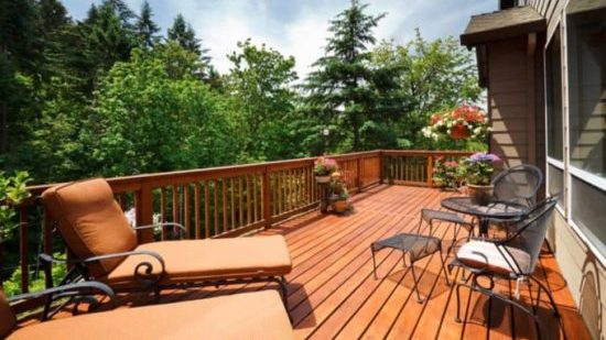 A wooden deck with lounge chairs, a table with chairs, potted plants, and flowers, surrounded by lush green trees under a partly cloudy sky.