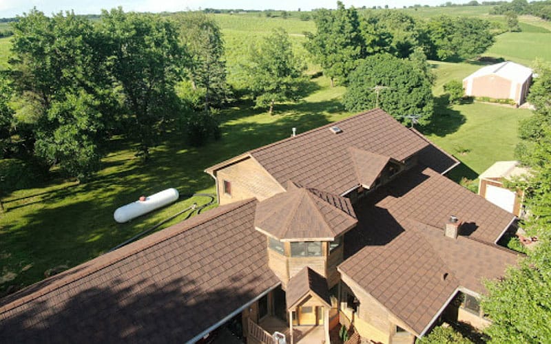 Aerial view of a large house with a brown roof surrounded by green trees and grass, a propane tank nearby, and a white barn in the background.