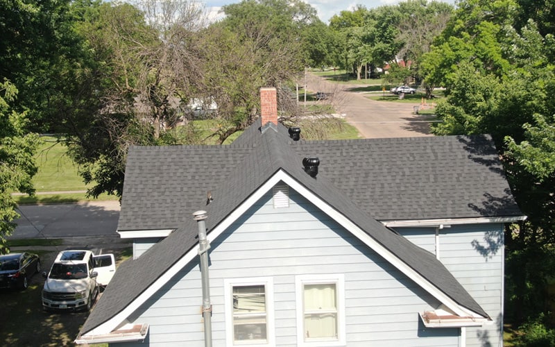 Aerial view of the roof of a light blue house with a brick chimney, surrounded by trees, parked cars, and a street visible in the background on a sunny day.