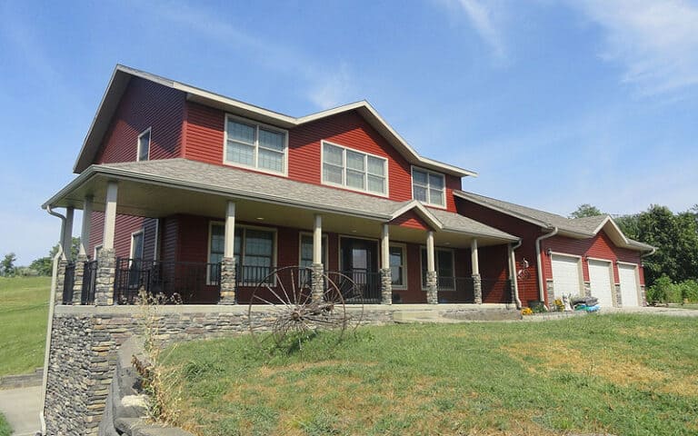 A two-story red house with white trim, a wraparound porch, and a three-car garage, sits on a grassy lawn with a stone retaining wall and an old wagon wheel decoration in front.
