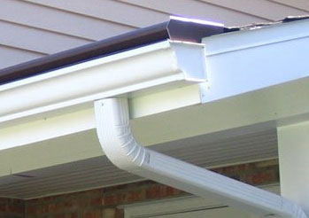White rain gutter system attached to the edge of a house roof with a downspout directing water downward; part of the roof and siding are also visible.