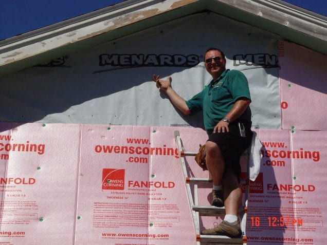 A man in sunglasses and a green shirt stands on a ladder, smiling and pointing at a house exterior covered with pink Owens Corning Fanfold insulation wrapping. The sun is shining, and the date on the photo is 16.