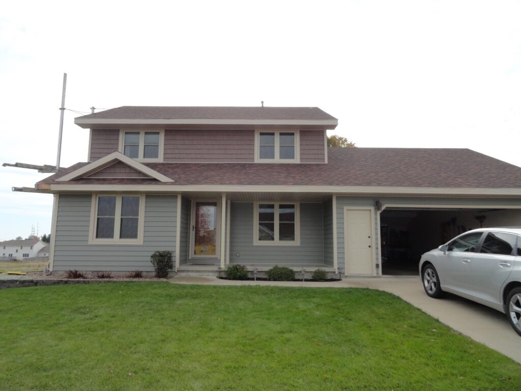 Two-story suburban house with gray siding, a brown roof, and attached garage. A white SUV is parked in the driveway, and the front yard has green grass and small shrubs.