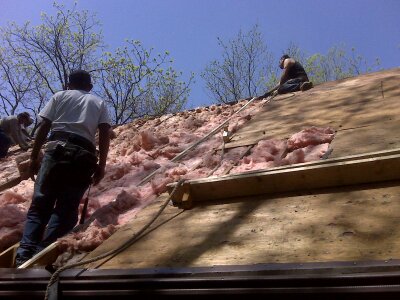 Several people work on a steep roof, installing pink insulation. They use safety ropes and harnesses, with trees and a clear blue sky visible in the background.