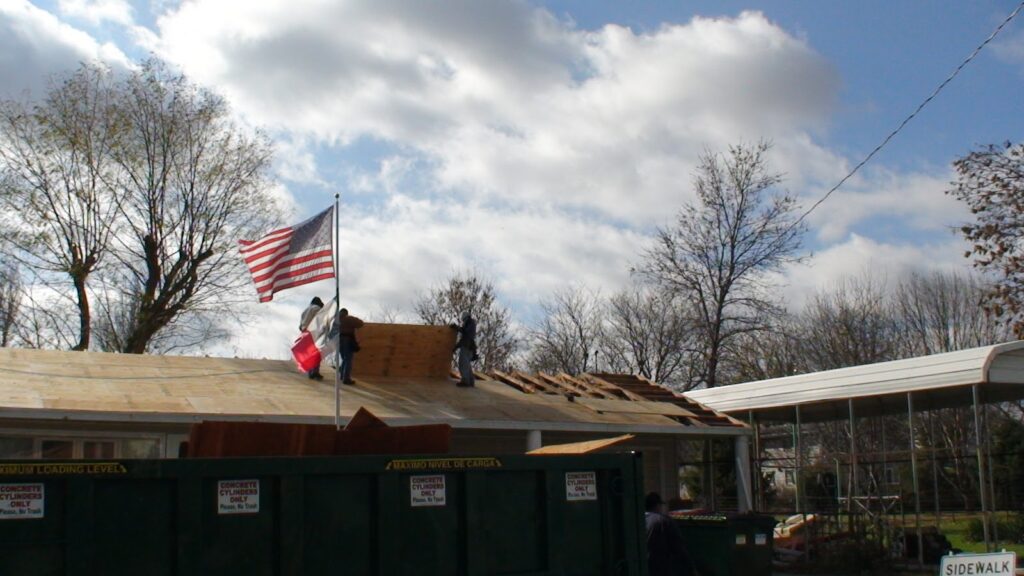 Two workers repair the roof of a house under partly cloudy skies. An American flag and another flag wave nearby. Trees without leaves surround the scene, and a large dumpster sits in front of the house.
