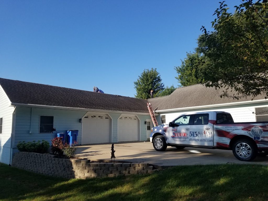 A light blue house with two garage doors, a ladder leaning against the roof, two workers on the roof, and a pickup truck with patriotic graphics and a phone number parked in the driveway on a sunny day.