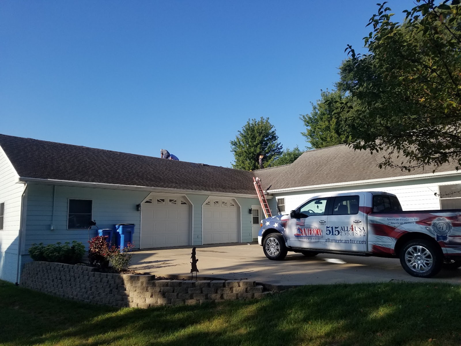 A light blue house with two garage doors, a ladder leaning against the roof, two workers on the roof, and a pickup truck with patriotic graphics and a phone number parked in the driveway on a sunny day.