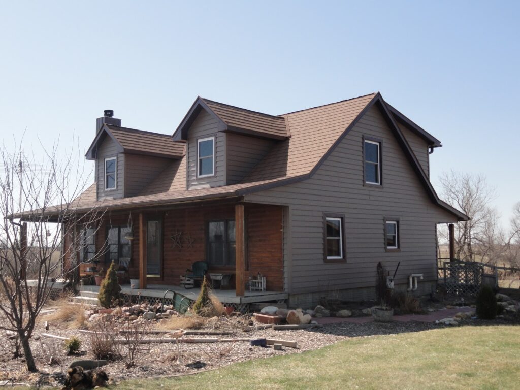 A two-story house with brown siding and a covered front porch sits in a yard with dry grass, small shrubs, and a rock garden under a clear blue sky.