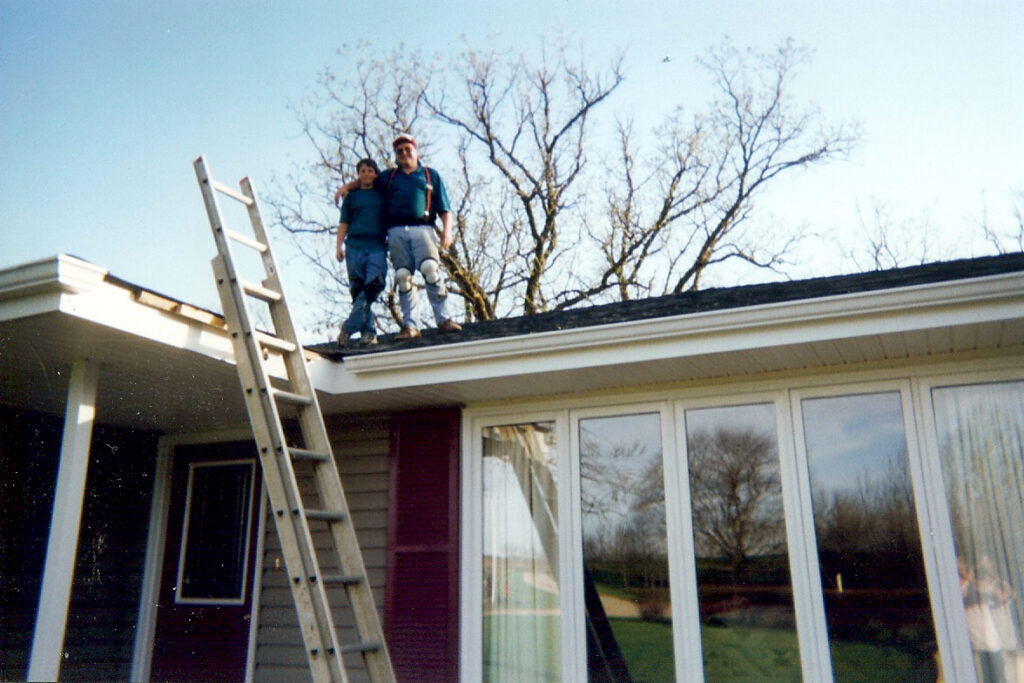 Two people stand on the roof of a house beside a ladder. Leafless trees are in the background and large windows reflect the outdoor scene. The sky is clear.