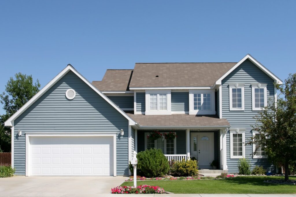 A two-story blue house with white trim, a double garage, front porch, manicured lawn, colorful flowerbeds, and a tree on the right, set against a clear blue sky.